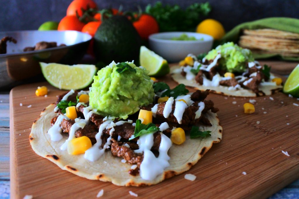 An angled image of two corn tortillas topped with pineapple-soy marinated beef, crema, guacamole, corn, and cheese on a wood board surrounded by fresh lime wedges, parsley, lemons, lime, a stack of corn tortillas, tomatoes, avocado, and beef in a pan.