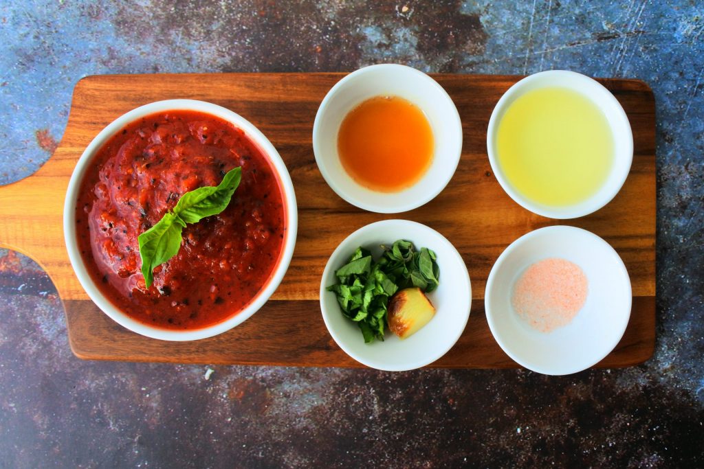 An over head image of a wooden plank with bowls of ingredients for homemade pizza sauce including crushed tomatoes, honey, olive oil, salt and fresh herbs