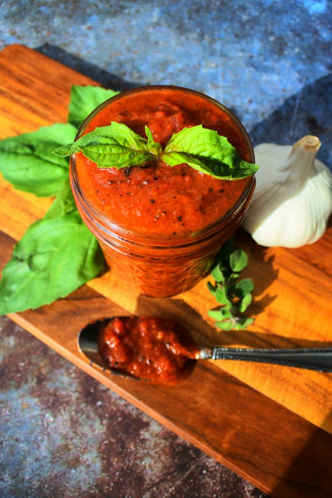 An overhead image of a jar of homemade pizza sauce on a wooden board with a spoon of tomato sauce, a head of garlic, a sprig of oregano and leaves of fresh basil around it