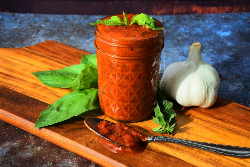 A head on image of a jar of homemade pizza sauce on a wooden board with a spoon of tomato sauce, a head of garlic, a sprig of oregano and leaves of fresh basil around it