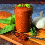 A head on image of a jar of homemade pizza sauce on a wooden board with a spoon of tomato sauce, a head of garlic, a sprig of oregano and leaves of fresh basil around it