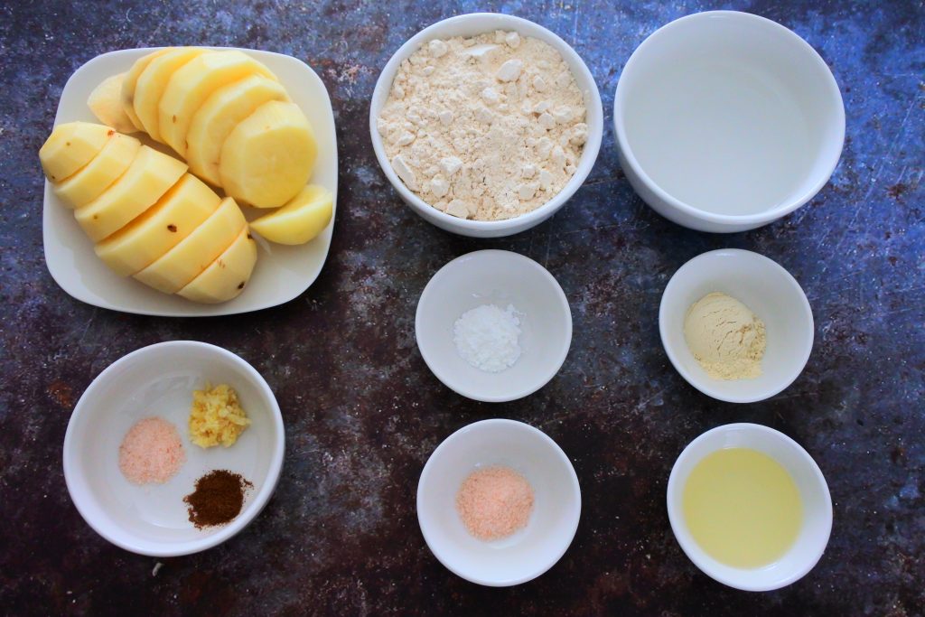 An overhead image of ingredients needed for potato samosas/aloo pies including: sliced potatoes, salt, garlic, ground geera (aka cumin), whole wheat flour, baking powder, salt, light olive oil, vital wheat gluten and water.