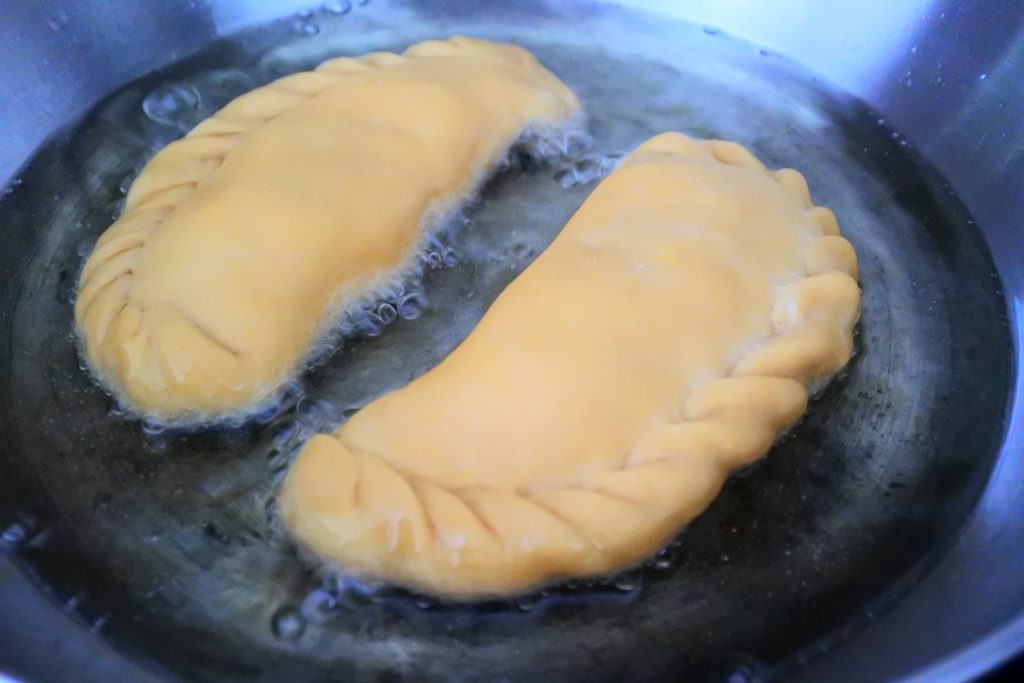 A close up image of potato samosas/aloo pies being added to a pan of shallow hot oil