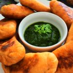 A close up image of a plate of potato samosas/aloo pies with a small bowl of cilantro dipping sauce in the middle and an adornment of fresh cilantro, lemon, garlic and potatoes in the background