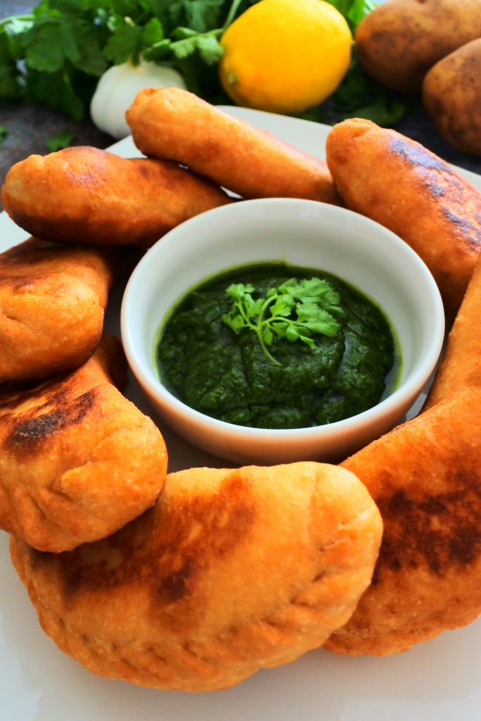A close up image of a plate of potato samosas/aloo pies with a small bowl of cilantro dipping sauce in the middle and an adornment of fresh cilantro, lemon, garlic and potatoes in the background