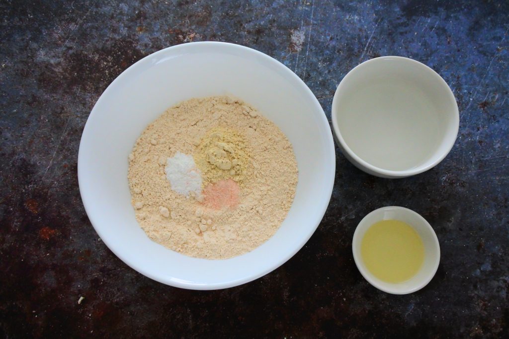 An overhead image of a bowl containing flour, baking powder, salt and vital wheat gluten, another bowl containing water and another bowl containing light olive oil