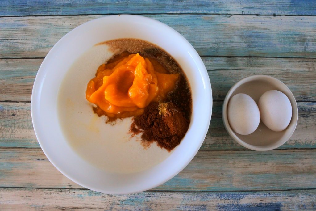 An overhead image of a large bowl with pumpkin puree, coconut milk, maple syrup and spices next to a smaller bowl containing two whole eggs