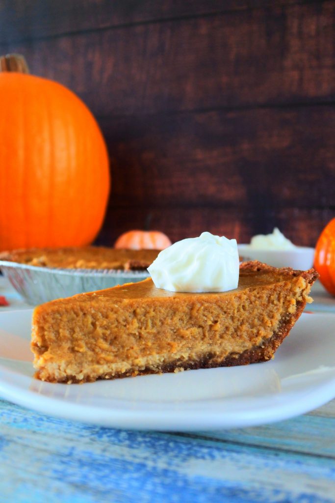 A vertical close up image of a slice of homemade pumpkin pie topped with coconut whipped cream on a white place with a large pumpkin in the background