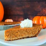 A close up image of a slice of pumpkin pie on a plate topped with a dollop of coconut whipped cream on a plate with a pie tin and pumpkins in the background