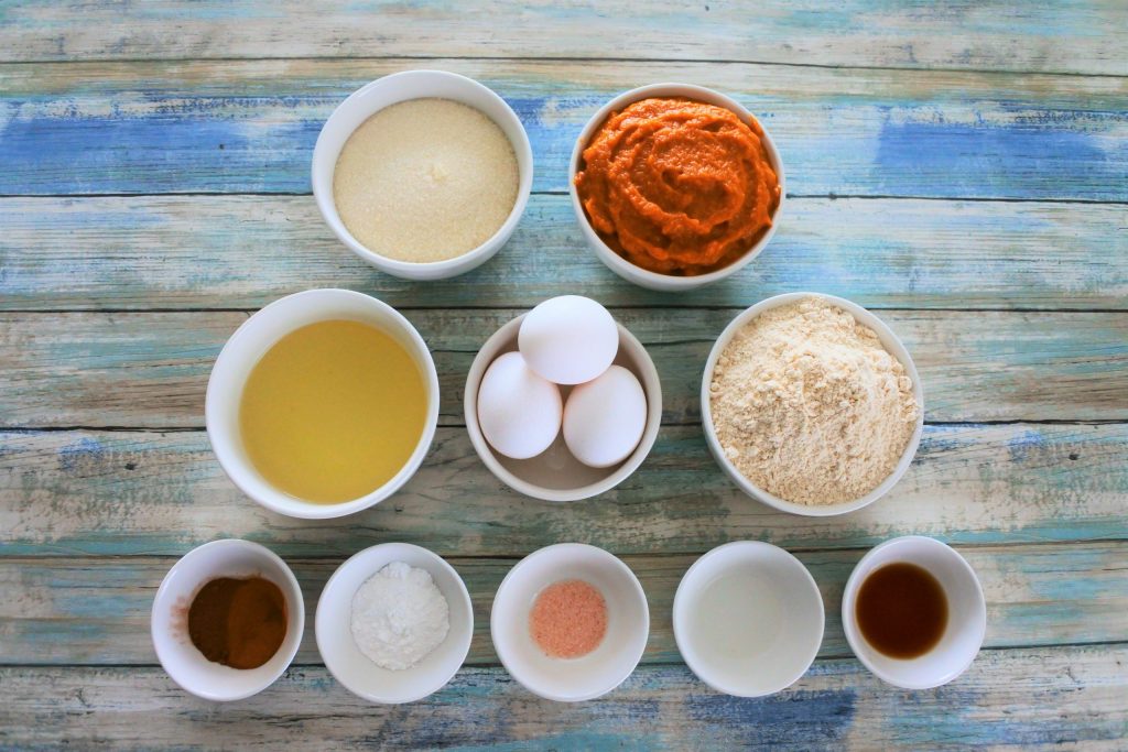 An overhead image of ingredients for a pumpkin spiced loaf including raw cane sugar, pumpkin puree, oil, eggs, whole wheat pastry flour, spices, baking powder, salt, and extracts.