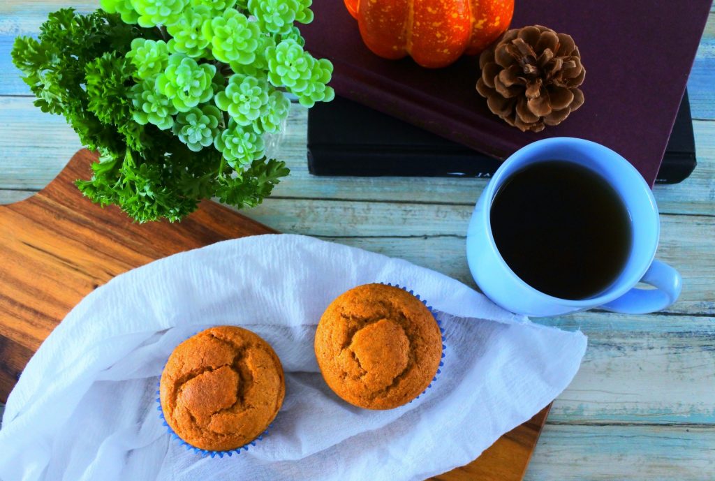 An overhead image of two pumpkin spice muffins on a white cloth on a board with another in the background along with a stack of books with a pumpkin on top, a cup of coffee and some greenery