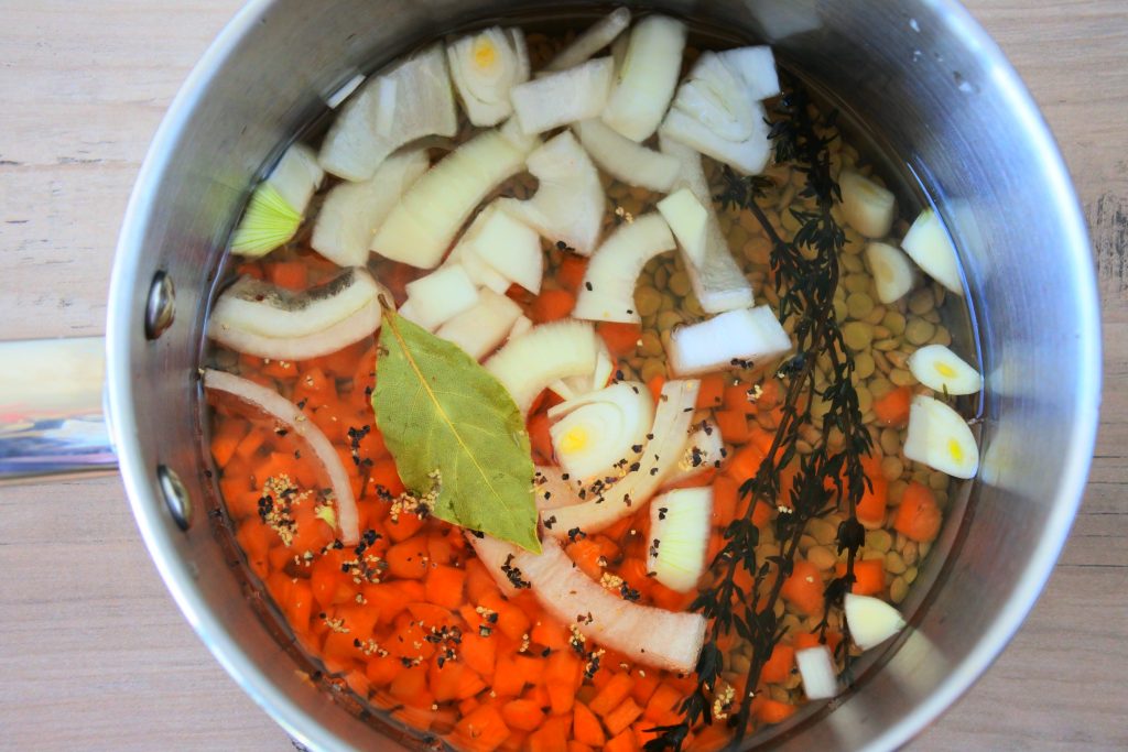 An overhead image of a pot filled with uncooked lentils, fine thyme, bay leaves, black pepper, diced carrots, cut onions, all of which are covered in water