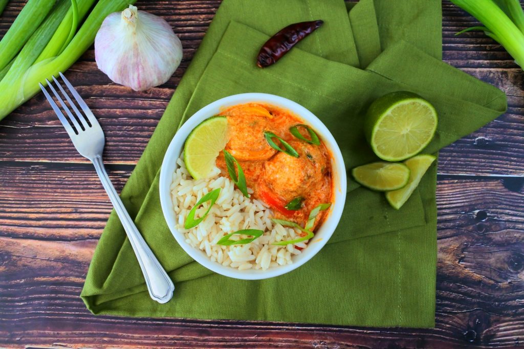 A overhead image of a bowl of coconut rice with red thai curry and thai chicken meatballs topped with sliced chives and a lime wedge on a green linen napkin with head of garlic, green onions, limes and a red pepper in the background
