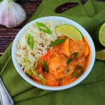 A head on angled image of a bowl of coconut rice with red thai curry and thai chicken meatballs topped with sliced chives and a lime wedge on a green linen napkin with head of garlic, green onions, limes and a red pepper in the background