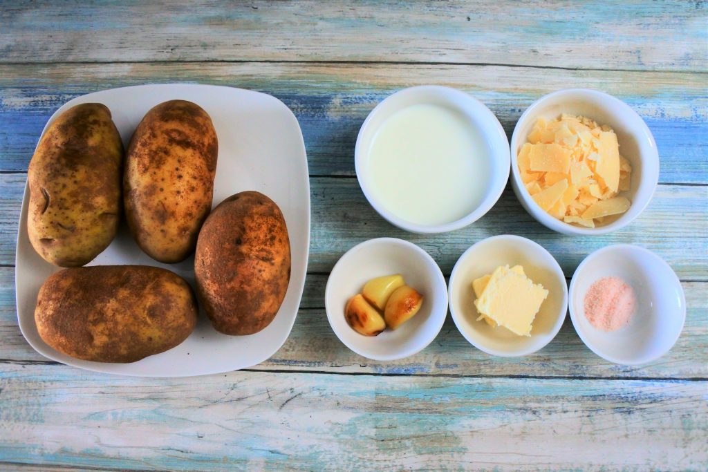 An overhead image of ingredients needed for roasted garlic and parmesan mashed potatoes including whole potatoes, milk, parmesan cheese, cloves of roasted garlic, butter and salt