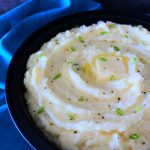 A vertical image of a bowl of roasted garlic parmesan mashed potatoes topped with a pat of butter, fresh cracked pepper and chives