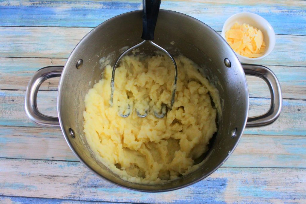 An overhead image of a pot with mashed potatoes and a potato masher with a small dish of parmesan on the side
