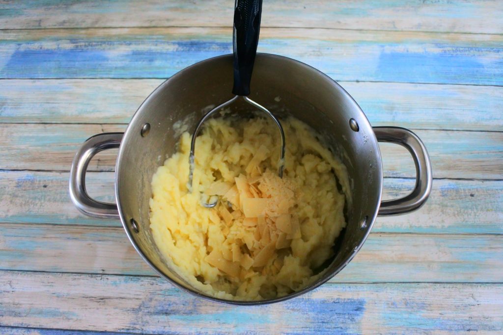An overhead image of a pot of mashed potatoes and a potato masher with shaved parmesan cheese added in