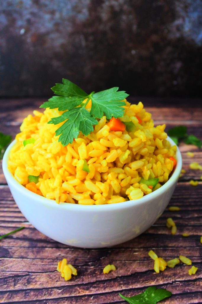 A close up vertical image of a bowl of saffron rice with diced carrots and green bell peppers mixed in and a garnish of fresh parsley