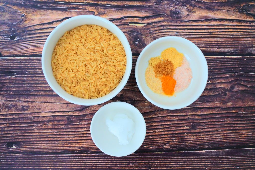 An overhead image of three bowls, one containing brown parboiled rice, one containing coconut oil and the third containing a blend of spices including powdered saffron, onion powder, garlic powder, salt, ginger powder and organic raw sugar
