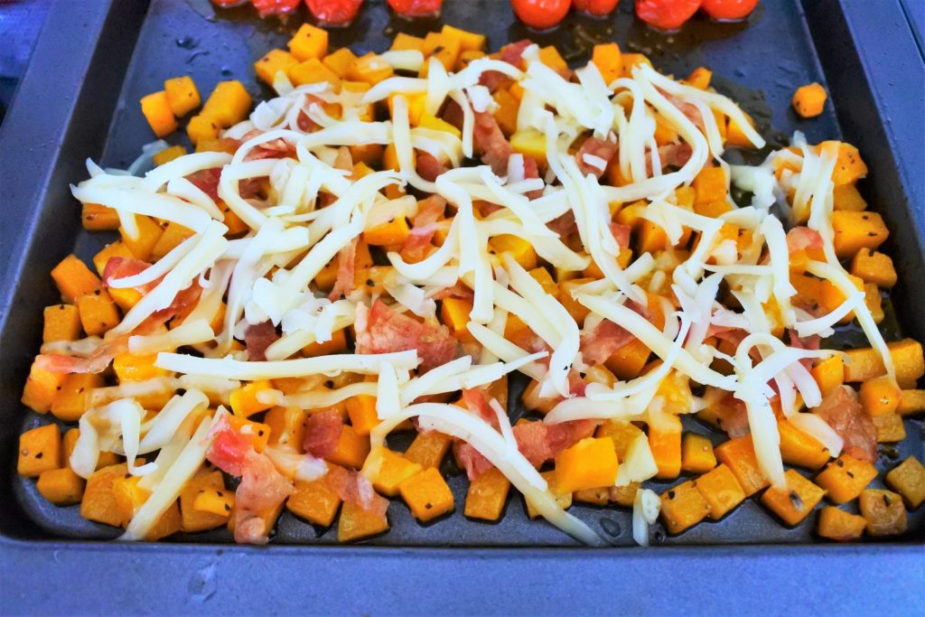 A close up image of shredded cheese and bacon being added to oven roasted butternut squash cubes