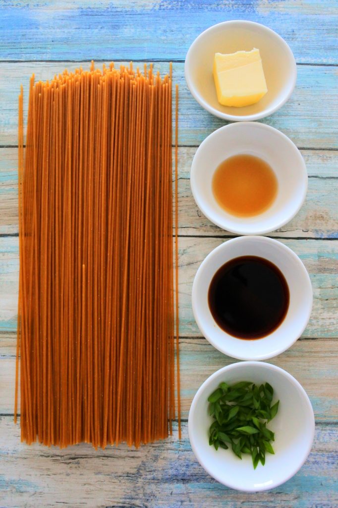 An overhead image of uncooked whole wheat spaghetti, butter, sesame oil, soy sauce and chopped chives