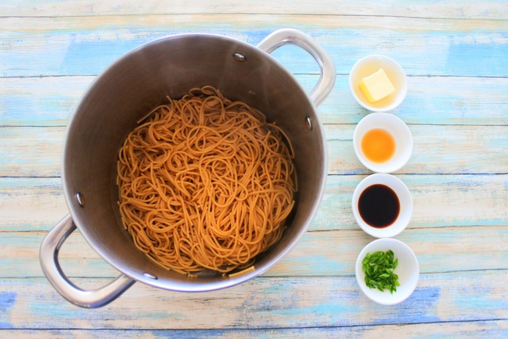 An overhead image of a pot of cooked whole wheat noodles with four smaller dishes next to it containing butter,sesame oil, soy sauce and chopped chives