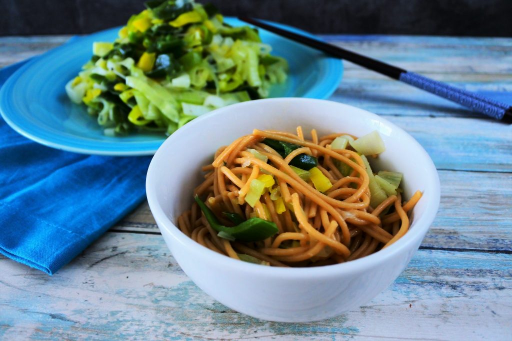 A head on image of a bowl of sesame noodles mixed with buttered leeks with a plate of buttered leeks on a blue linen napkin with a pair of chopsticks in the background