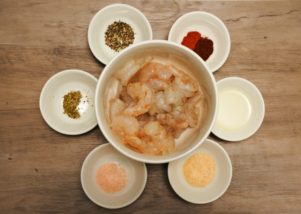 An overhead image of a bowl of peeled and deveined shrimp surrounded by smaller dishes of spices including salt, black pepper, light olive oil, dried oregano, garlic powder, smoked paprika, and chipotle powder