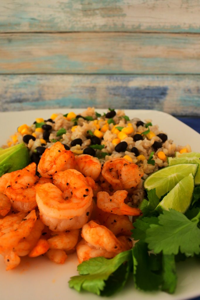 A tilted angle image of a plate of chipotle fried shrimp served with a corn and black bean rice and garnished with fresh parsley and lime wedges