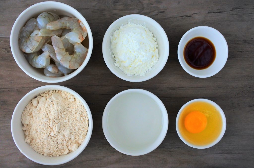 An overhead image of six bowls, each containing a single ingredient: shrimp, whole wheat pastry flour, potato starch, ice water, soy sauce, and an egg