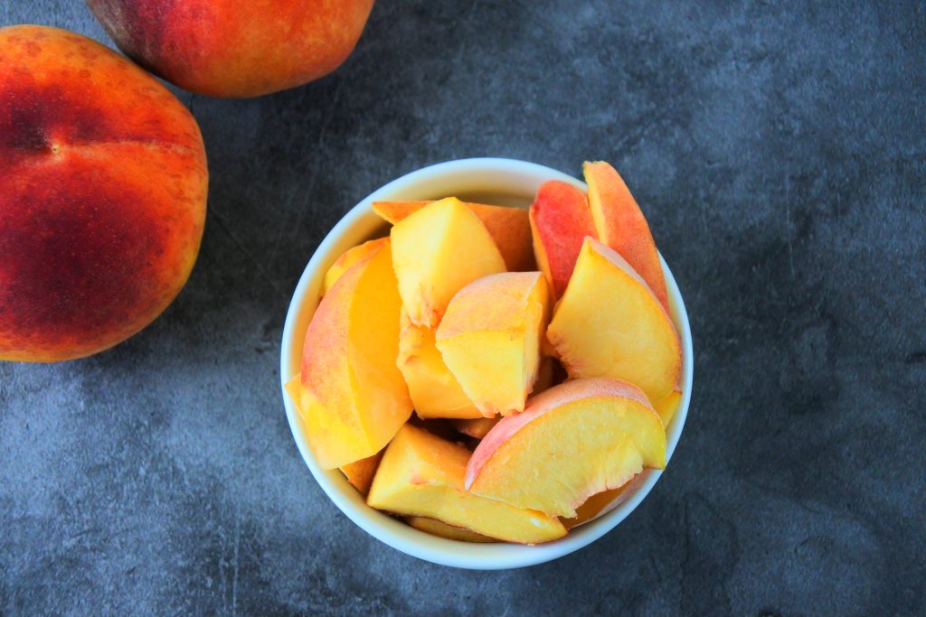 An overhead image of a bowl of frozen peach chunks with whole fresh peaches in the top left corner