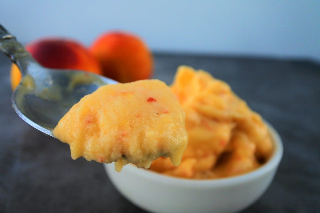 A close up image of a spoonful of one ingredient peach sorbet with a bowl of peach sorbet and whole fresh peaches in the background