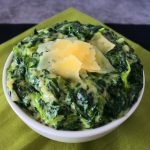 An angled image of a bowl of creamed spinach topped with shaved parmesan on a green and black linen napkin.