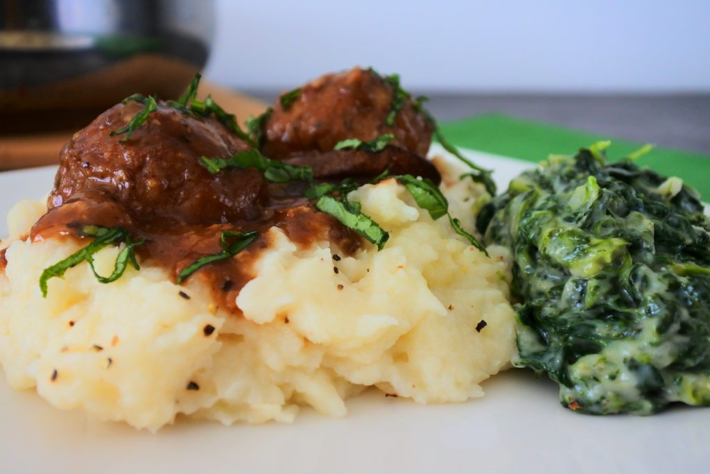 A close up image of mashed cauliflower topped with salisbury meatballs and served with a side of creamed spinach