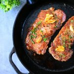 An overhead image of seared steak topped with garlic and thyme in a cast iron skillet with a cropping of herbs in the top left edge