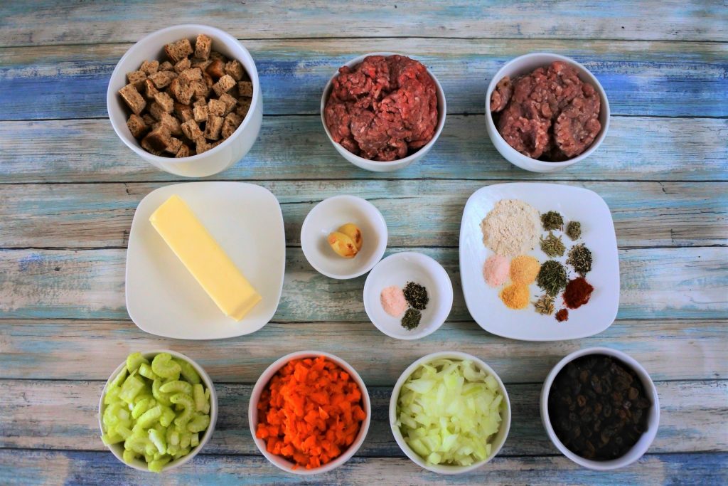 An overhead image of plates and bowls of ingredients including croutons, ground beef, ground pork, a stick of butter, roasted garlic, spices, celery, carrots, onions, and raisins