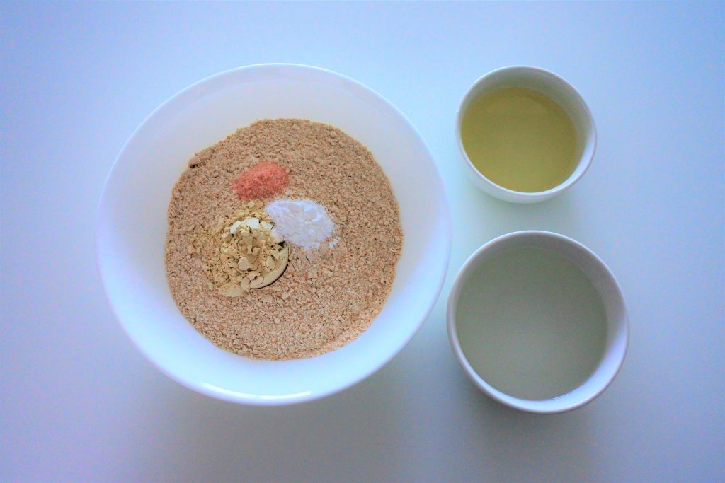 And overhead image of a bowl containing white whole wheat flour, vital wheat gluten, salt and baking power and two bowls to the right containing oil and water