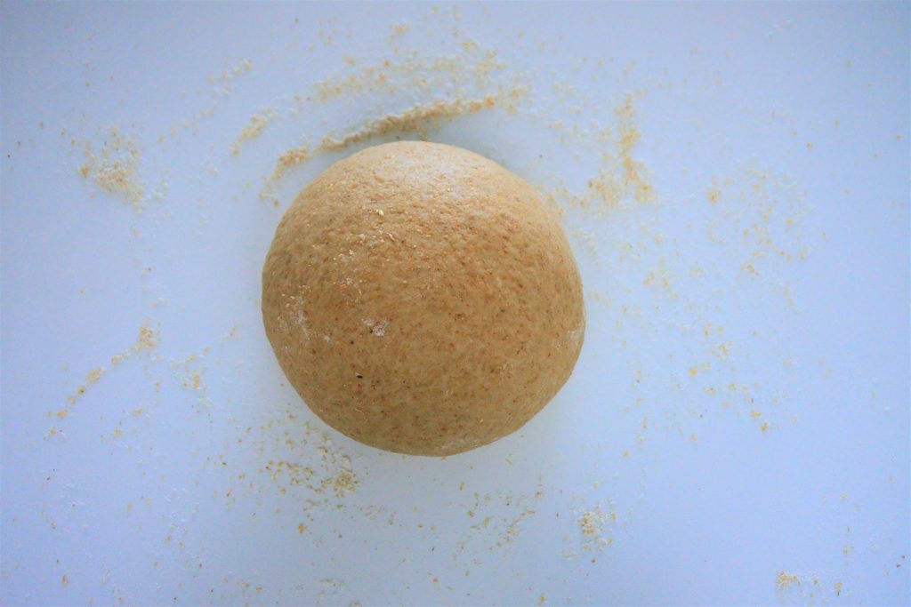 An overhead image of a ball of whole wheat tortilla dough surrounded by a scattered sprinkle of flour
