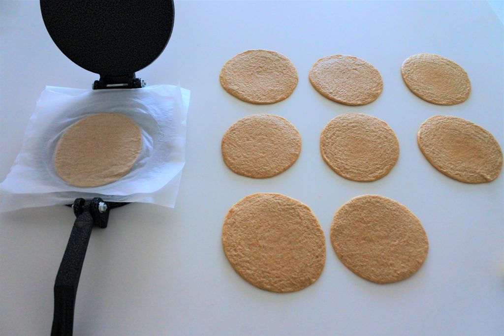 An angled image of rounds pressed tortilla dough on a white countertop with one dough round between two pieces of parchment paper that is sitting in an opened cast iron tortilla press