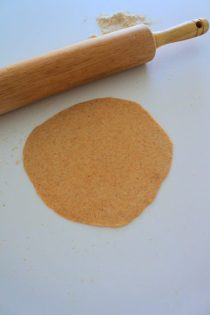 Overhead image of a round of tortilla dough rolled out against a white countertop with a rolling pin in the top left corner